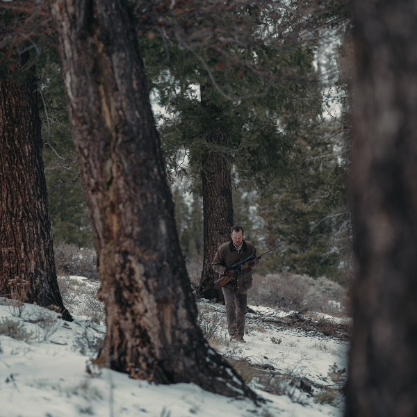Person walking through a snowy forest with trees and a rifle.