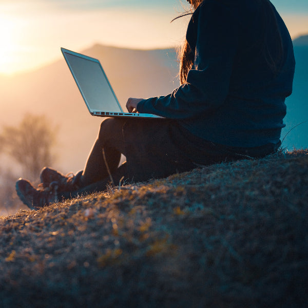 Person using a laptop in a scenic outdoor setting with mountains and sunset.