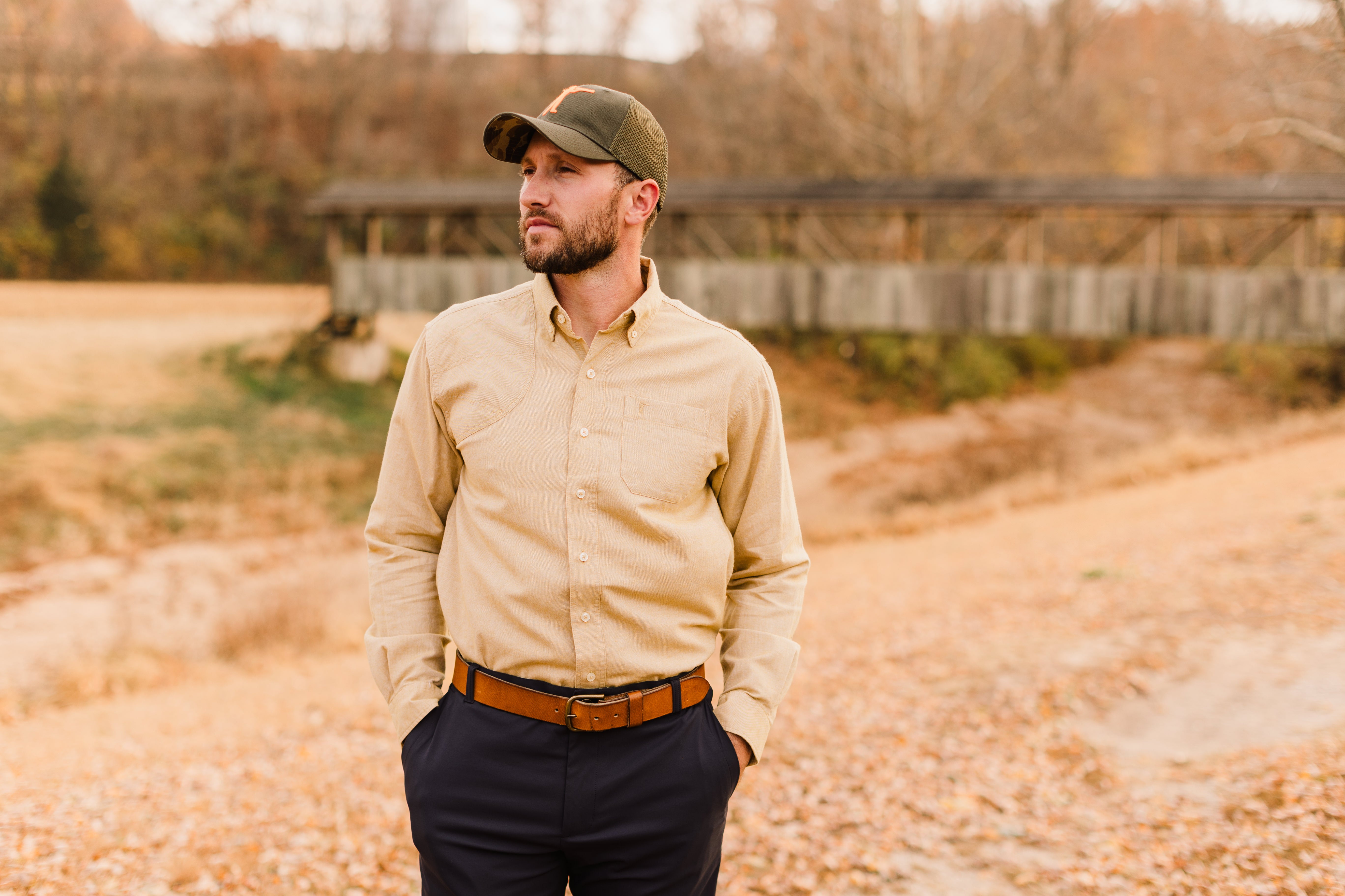 Man wearing a beige shirt, dark pants, and a cap standing outdoors with a blurred background