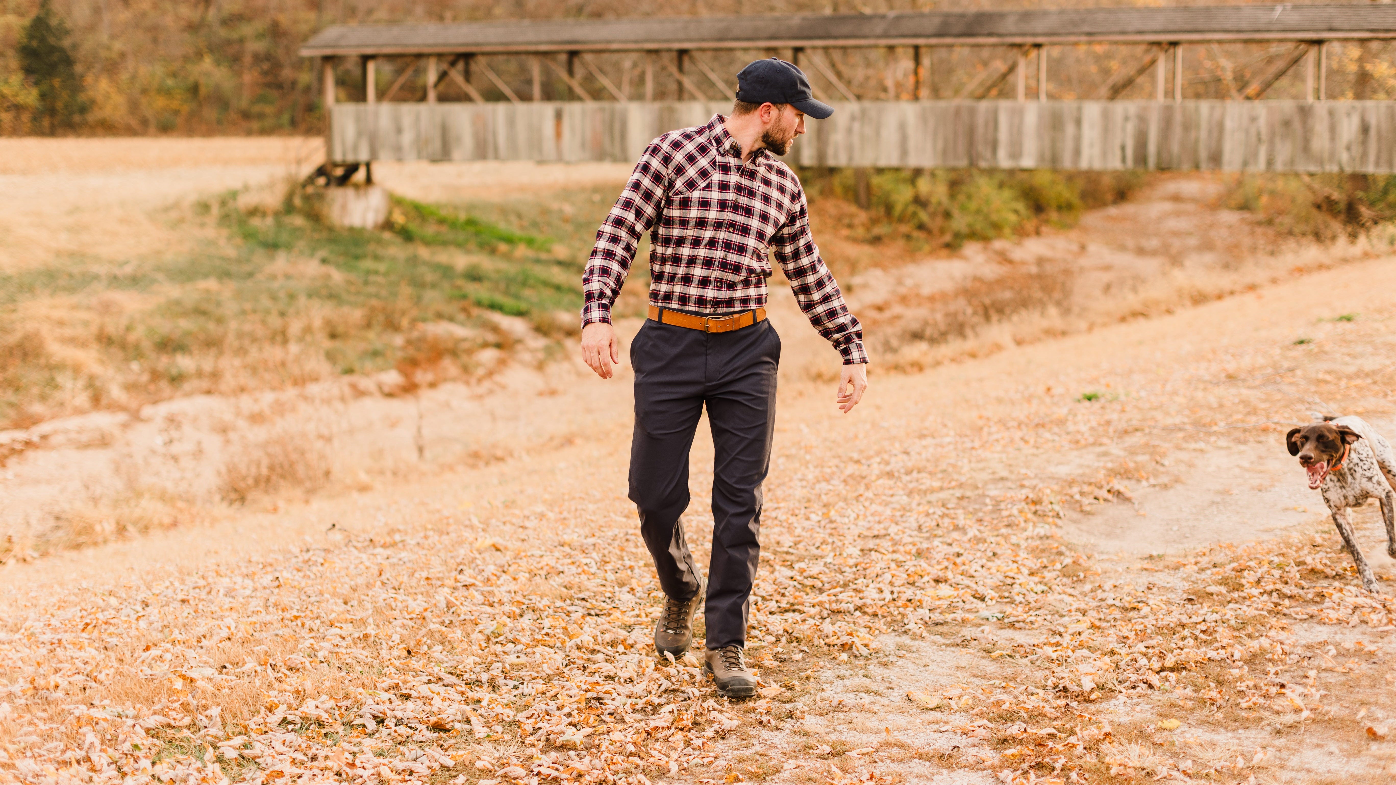 Man in checkered shirt and cap walking with a dog in a field.