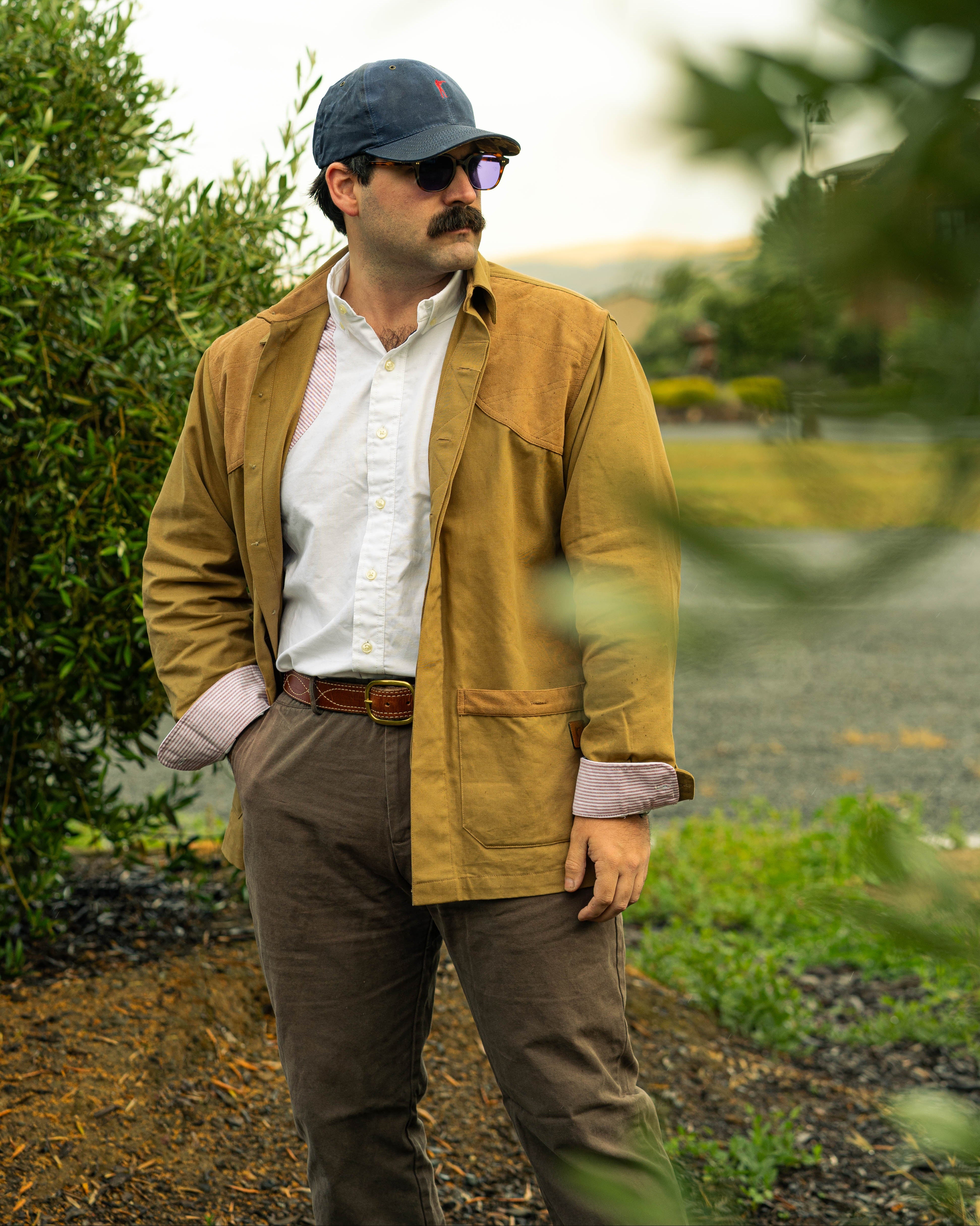 Man in a mustard jacket and cap standing outdoors with trees and a path in the background