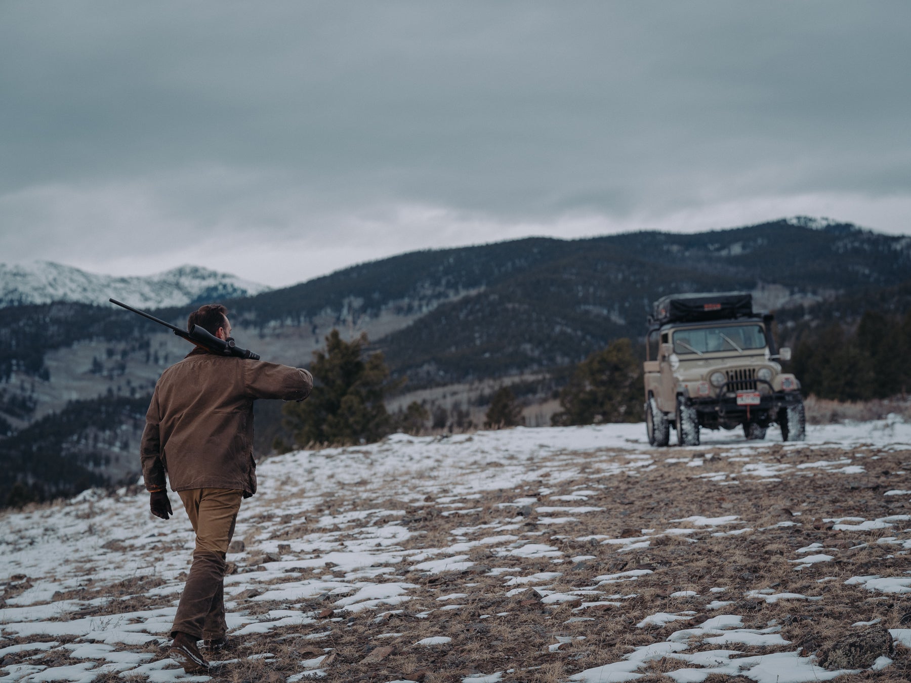 Man with a rifle standing in a snowy field with a truck in the background