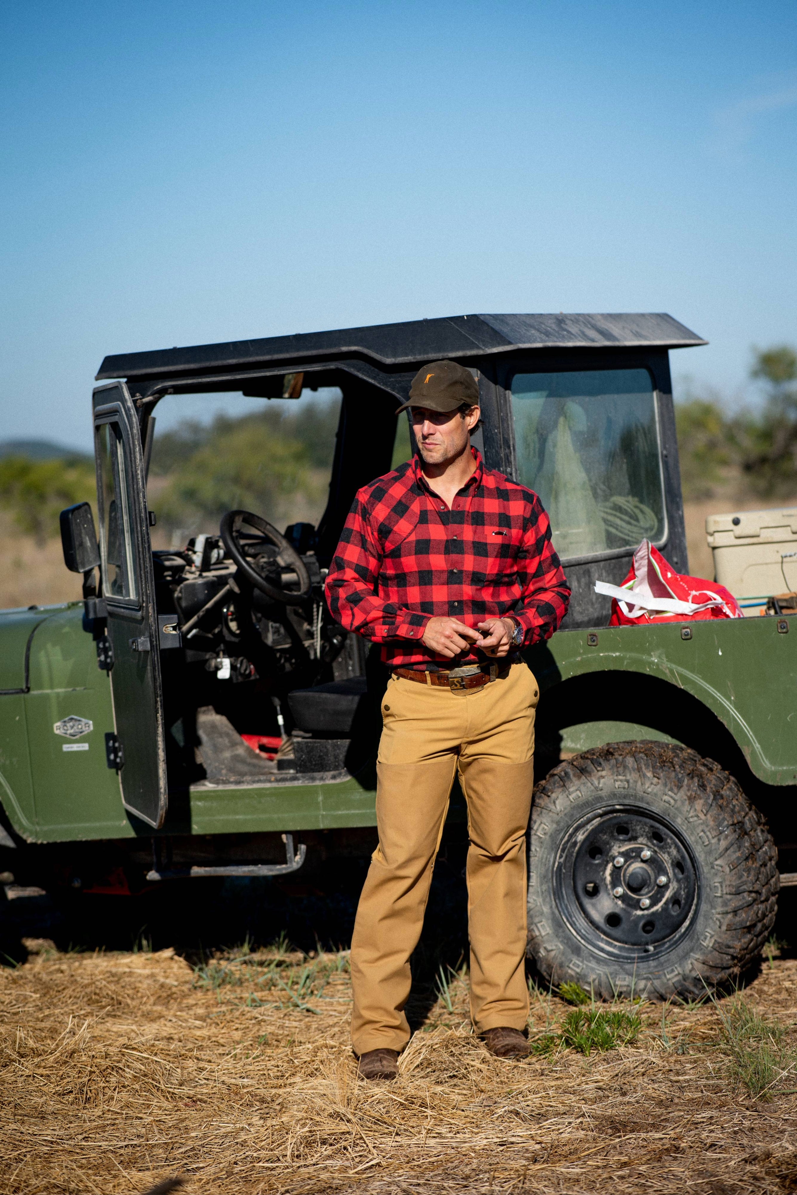 Man in a red plaid shirt and tan pants standing next to a green off-road vehicle in a natural setting.