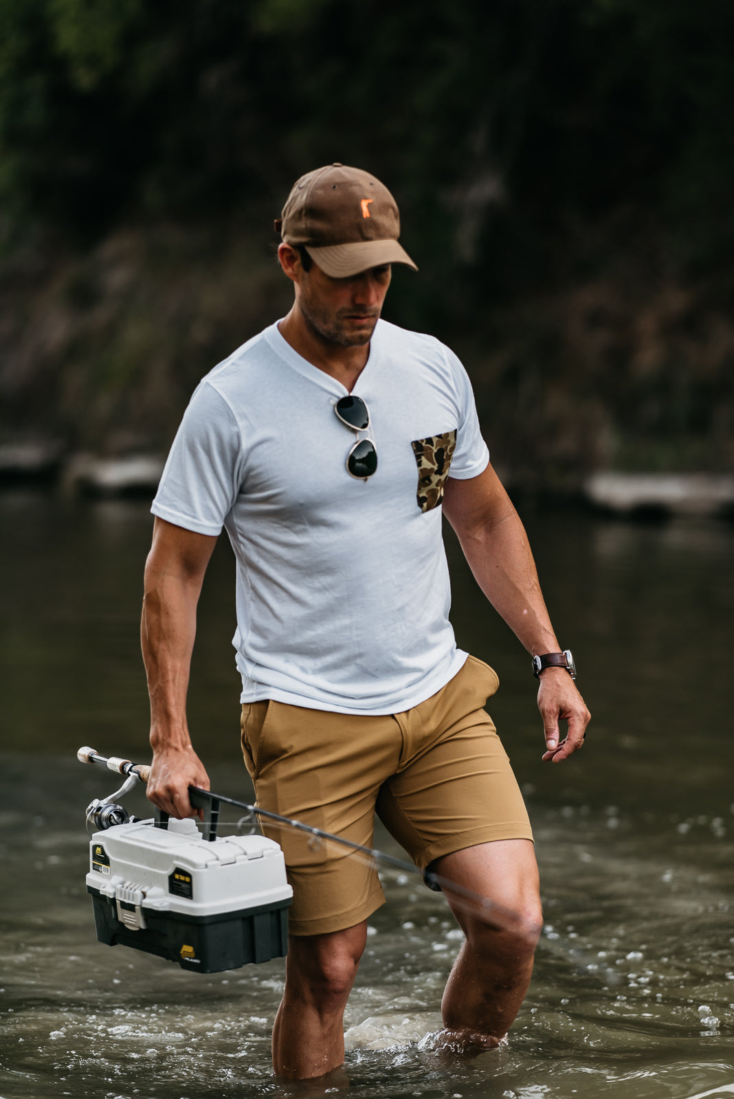 Man walking through water carrying a cooler, wearing a white t-shirt and beige shorts.