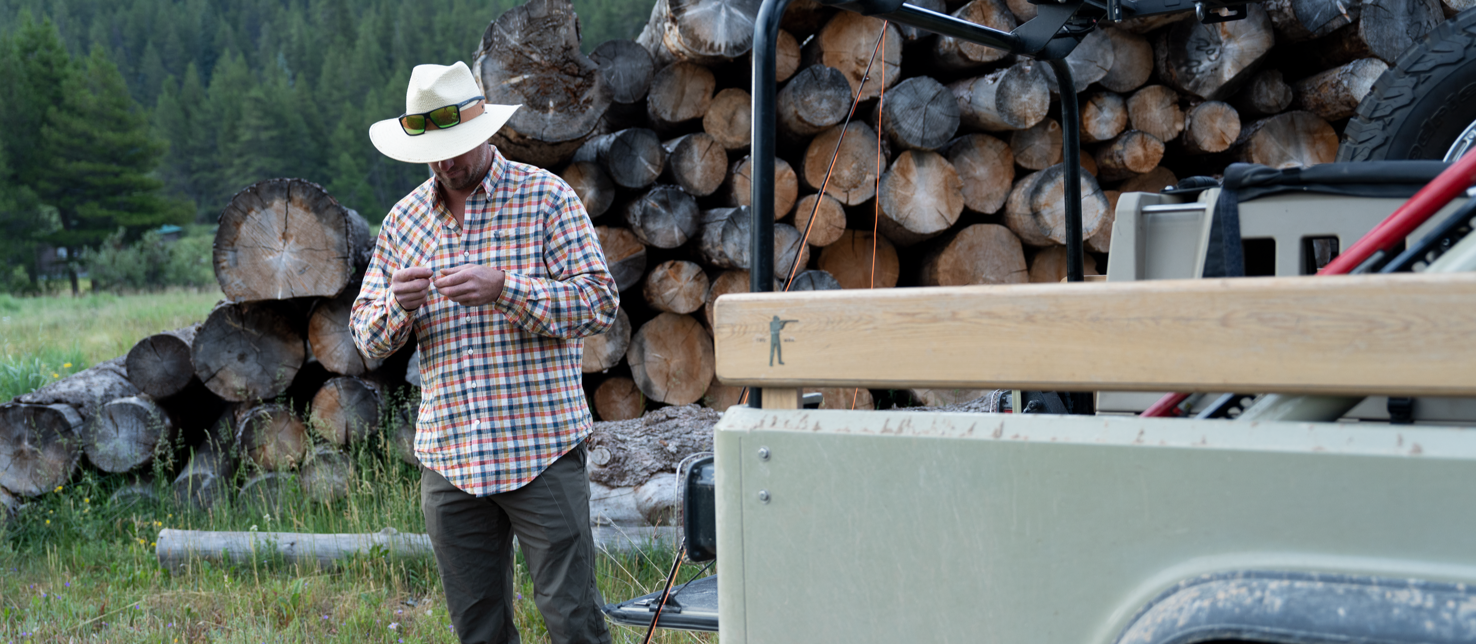 Man in a cowboy hat standing next to a truck with wood logs in a forest setting