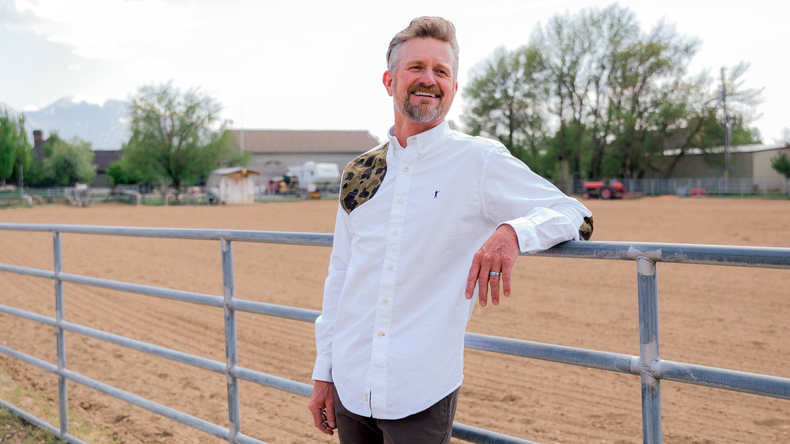 Man in a white chef's coat standing in an outdoor setting with a fence and open space.