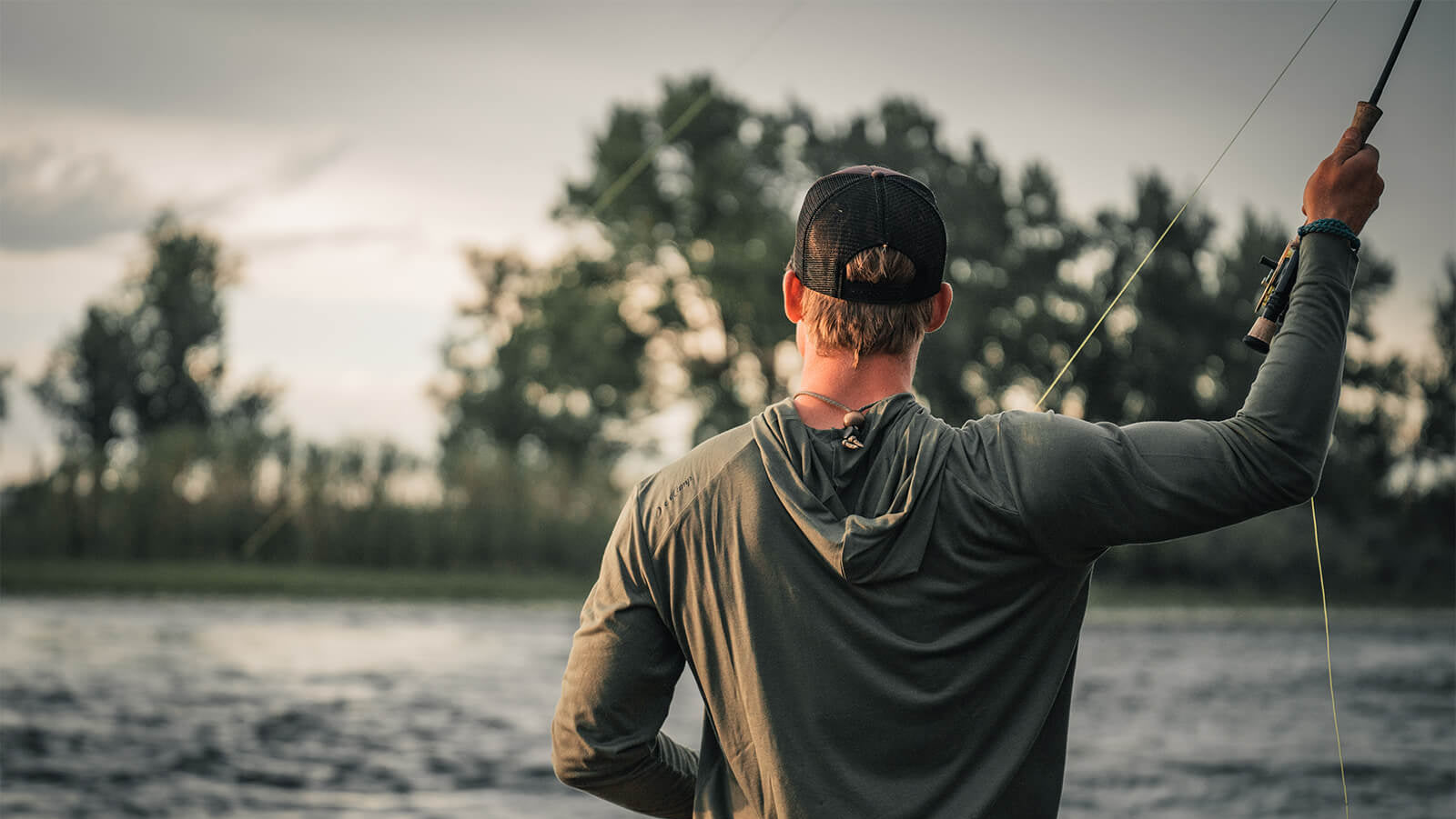 Man fly fishing by a body of water with trees in the background