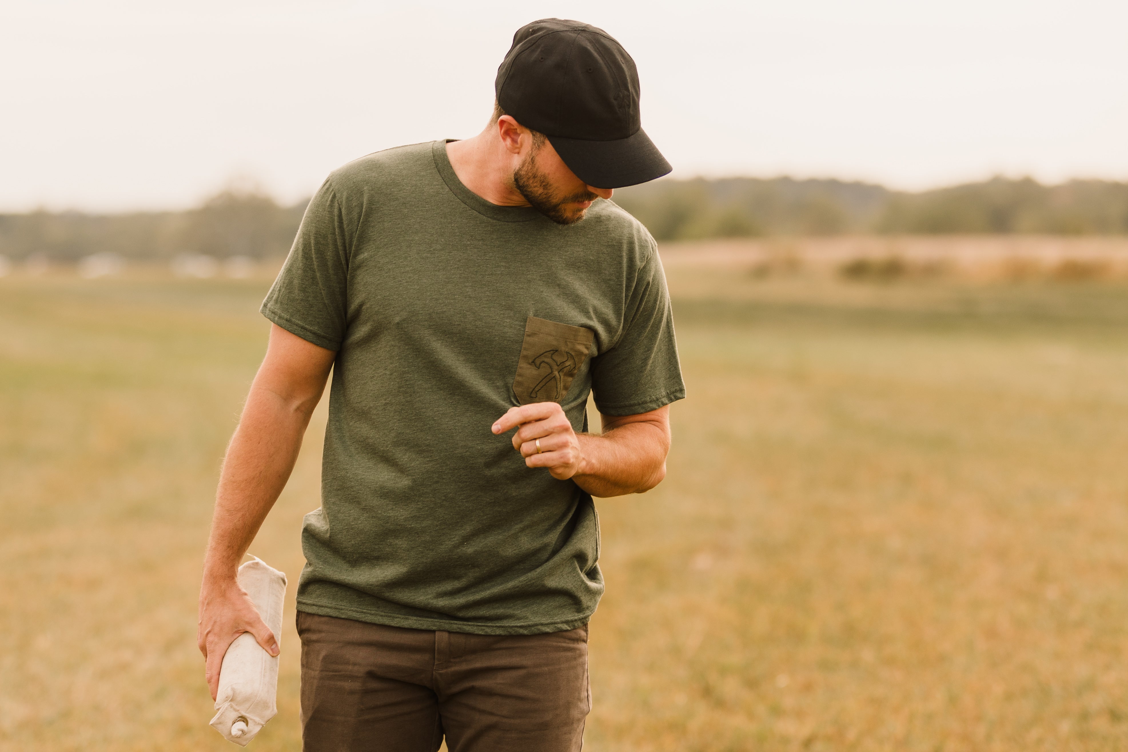 Man in a green t-shirt and black cap standing in a field holding a bottle.