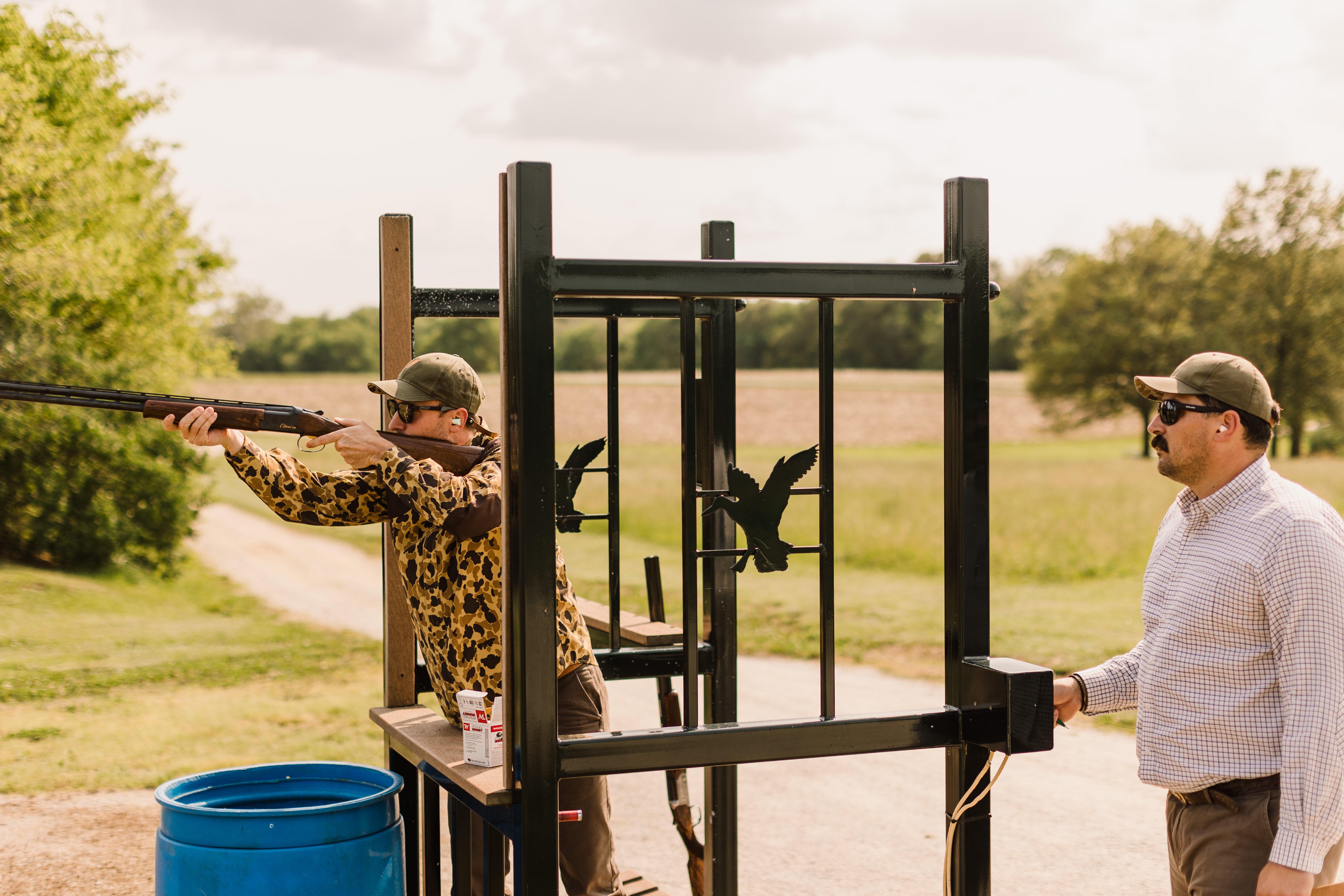 Two men at a shooting range with one aiming a shotgun