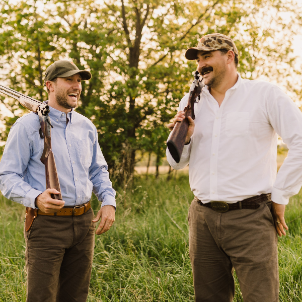 Two men in a field holding rifles, surrounded by trees and greenery.