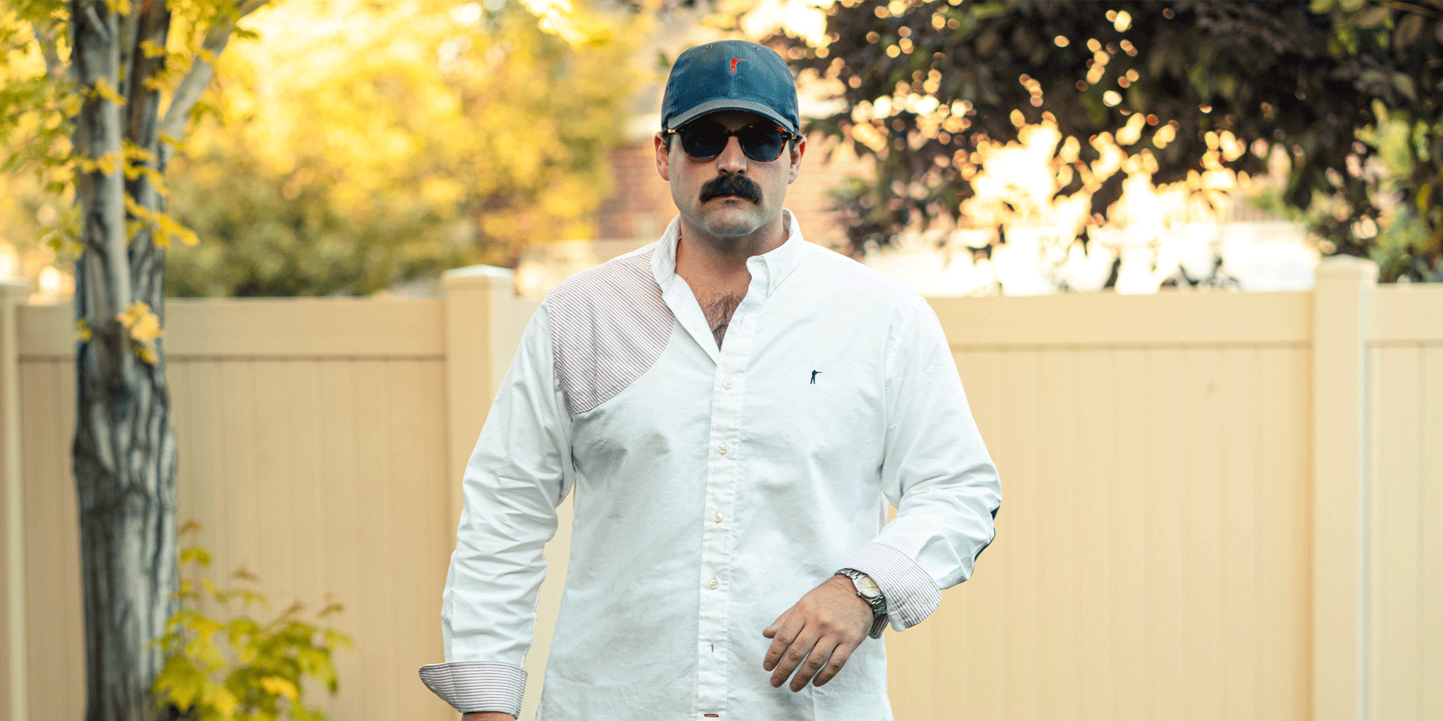 Man wearing a white shirt and blue cap outdoors with trees and fence in the background